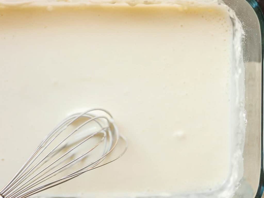 A whisk stirring a tempura batter in a glass baking dish, suggesting a mixture being prepared. The liquid is smooth and evenly distributed across the dish.